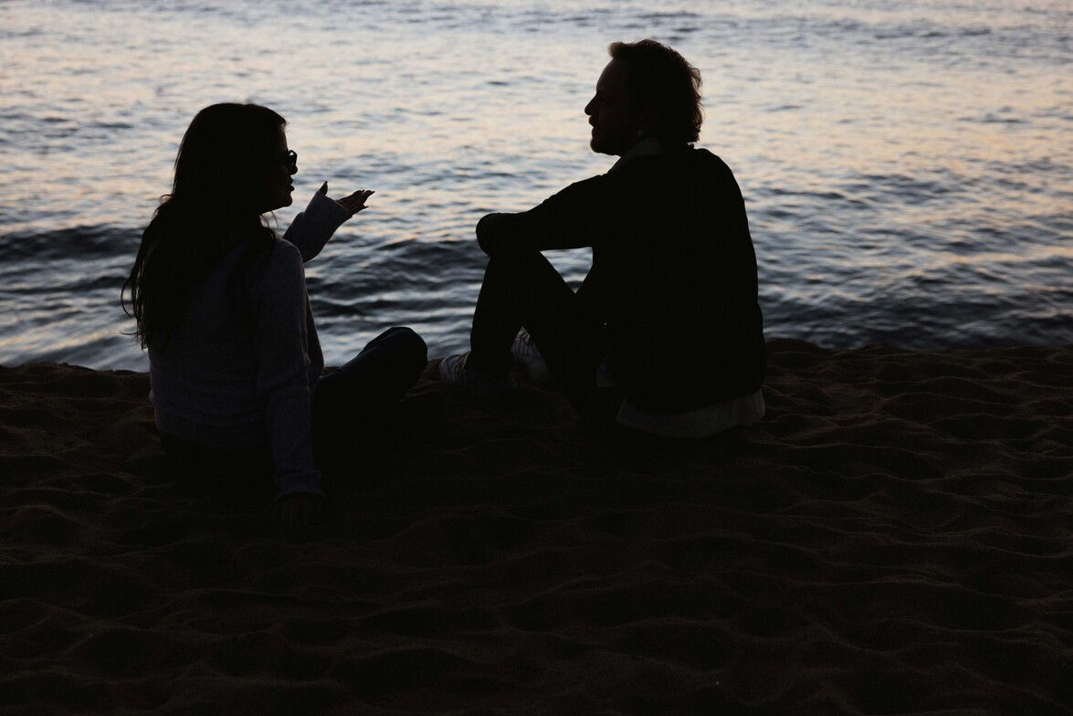 Two people sitting together beside the water at dusk, facing one another in conversation.